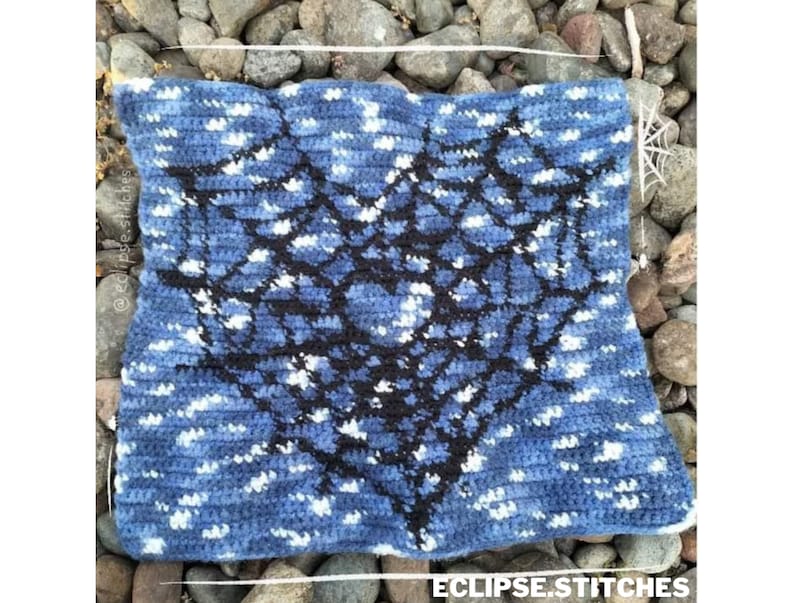 Op de afbeelding: Een blauw en wit gecrochet vierkant met een zwart geometrisch ontwerp in het midden. Het ontwerp lijkt op een spinnenweb.