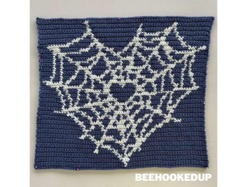 Op de afbeelding: Een blauw gehaakt vierkant met een wit gehaakt spinnenwebontwerp in het midden. Het vierkant is vastgemaakt aan een wit oppervlak. De tekst "BEEHOOKEDUP" is onder het vierkant gedrukt.