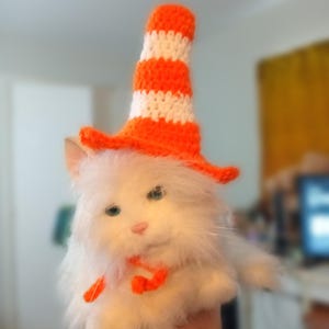 May include: A white plush toy cat wearing an orange and white crocheted traffic cone hat. The cat has blue eyes and a pink nose.