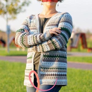 May include: A long, open-front crochet cardigan in a neutral color palette of beige, gray, and brown, with green accents. The handmade sweater features a striped pattern and open weave design, perfect for layering.