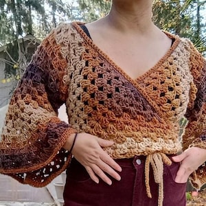 May include: A crocheted wrap top with a V-neck and long sleeves. The top is made in a brown, beige, and cream colour gradient. The top is tied at the waist with a long tie.