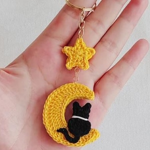 May include: A handmade yellow crochet keychain featuring a star and a crescent moon with a black cat silhouette. The keychain is attached to a gold-coloured metal ring, perfect for keys or bags.