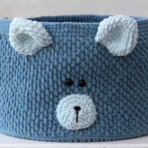 May include: Blue knitted storage basket with a bear face design. The basket has two large blue ears and a brown nose.