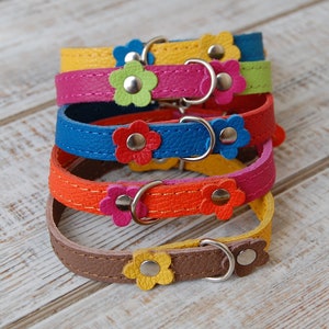 Può includere: Una pila di collari colorati per animali domestici. Ogni collare è di un colore diverso, tra cui giallo, rosa, blu, arancione e marrone. Ogni collare ha un abbellimento a forma di fiore e una fibbia in metallo argentato.