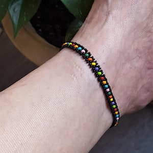 Op de afbeelding: Een kralenarmband met een zwarte basis en kleurrijke kralen in een regenboogpatroon. De armband wordt om een pols gedragen. De kralen zijn klein en rond, waardoor een textuur ontstaat. De armband is ongeveer 18 cm lang.