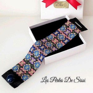 Puede incluir: Una pulsera de cuentas coloridas con un patrón floral geométrico en azul, rosa y verde, presentada en una caja de regalo blanca con una cinta roja. La pulsera tiene una base negra y un cierre de piedra azul. El texto "Les Perles De Sissi" es visible.