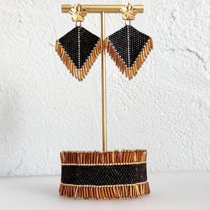 May include: A pair of diamond-shaped beaded earrings and a matching wide cuff bracelet. The jewelry features a black and metallic gold color scheme with intricate beadwork and dangling gold fringe accents, displayed on a gold metal stand.