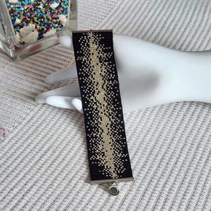 May include: A wide cuff bracelet crafted from small, uniform beads in a black and metallic gold color scheme. The design features a central vertical column of gold beads that transitions into a scattered, pixelated pattern against a black background.