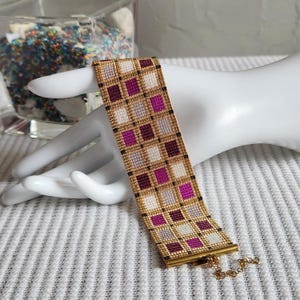 Op de afbeelding: Een brede kralenarmband met een rasterpatroon van kleine vierkantjes in tinten magenta, donkerbordeaux, lichtgrijs en wit, omlijnd met goudkleurige kralen en zwarte accenten, afgewerkt met een goudkleurige metalen staaf en een kettingsluiting.