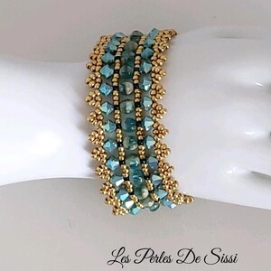 May include: A wide beaded bracelet featuring rows of metallic gold seed beads, faceted light blue crystal bicones, and central marbled teal round beads. The design includes intricate gold beadwork edges and the text Les Perles De Sissi.