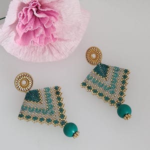 May include: A pair of handcrafted earrings with a geometric design. The earrings feature a teal, gray, and gold beaded pattern, with a teal bead at the bottom. The earrings have a gold-colored circular stud.