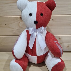 Pu&ograve; includere: Un orsacchiotto di peluche fatto a mano con un motivo a scacchi bianco e rosso. L'orso ha occhi di bottone neri e un fiocco di raso bianco.