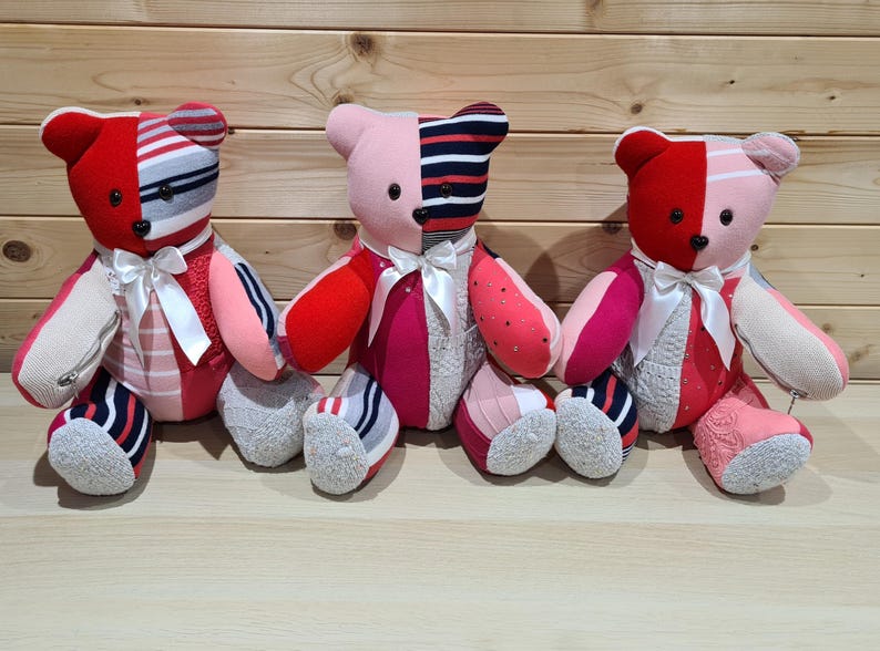 May include: Three handmade teddy bears with patchwork designs. Each bear features a mix of red, pink, and white fabrics, with striped and patterned details. They have white ribbon bows and gray sock feet.