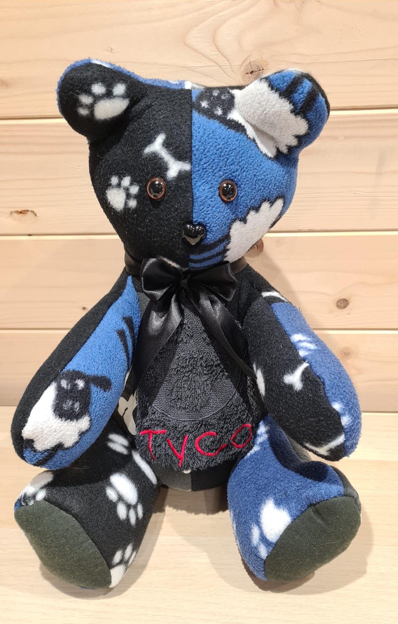 Pu&ograve; includere: Un orsacchiotto di pezza patchwork nero e blu con impronte di zampe bianche e un fiocco nero. L'orsacchiotto &egrave; seduto su una superficie di legno e ha il nome "Tyco" ricamato sul petto.