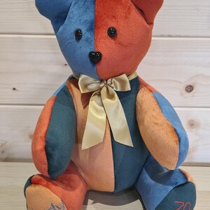 Pu&ograve; includere: Un orsacchiotto di peluche patchwork colorato con un fiocco dorato. L'orso &egrave; seduto su una superficie di legno. Le zampe dell'orso sono ricamate con i nomi "Wendy" e "70".