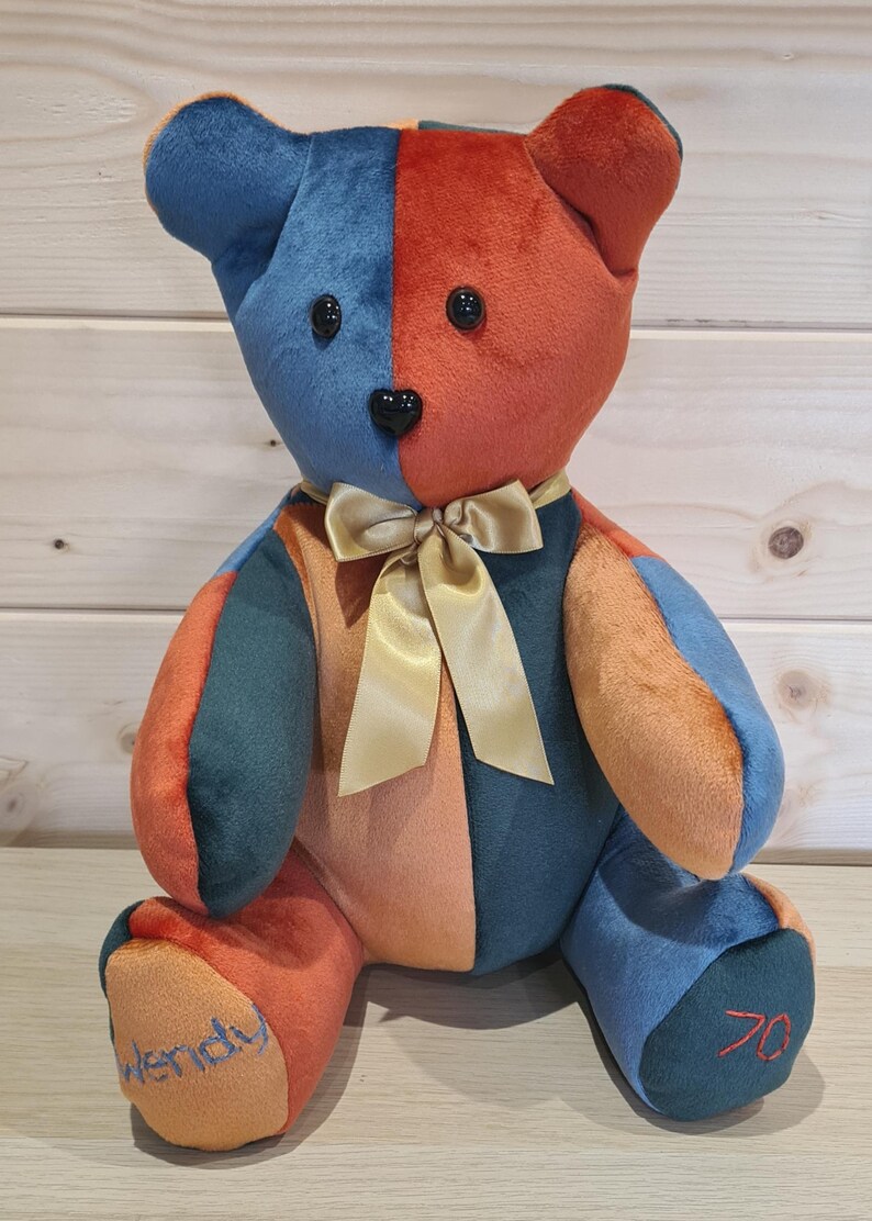 Pu&ograve; includere: Un orsacchiotto di peluche patchwork colorato con un fiocco dorato. L'orso &egrave; seduto su una superficie di legno. Le zampe dell'orso sono ricamate con i nomi "Wendy" e "70".