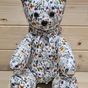Könnte beinhalten: Ein handgefertigter Teddybär mit einem Blumenmuster in Weiß, Orange, Lila und Grün. Der Bär hat schwarze Knopfaugen und eine schwarze Nase. Er trägt ein Hemd mit Kragen und dem gleichen Blumenmuster. Der Bär sitzt auf einer Holzoberfläche.