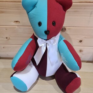 May include: A handmade teddy bear with a split design of teal and burgundy fabric. The bear has black button eyes, a black nose, and a white bow tie. The bear's arms and legs are a mix of colors, and the bear is sitting upright.