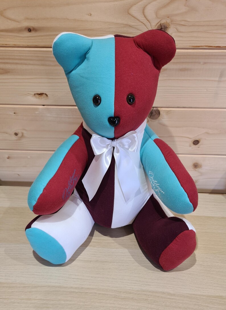 May include: A handmade teddy bear with a split design of teal and burgundy fabric. The bear has black button eyes, a black nose, and a white bow tie. The bear's arms and legs are a mix of colors, and the bear is sitting upright.