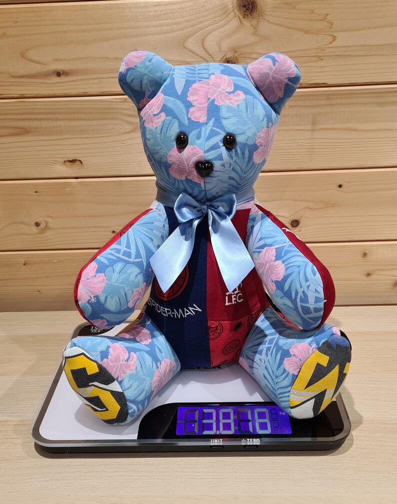 May include: A handmade teddy bear with blue fabric featuring pink floral patterns. The bear has a blue bow, and its arms and legs are red and blue. The bear is sitting on a digital scale.