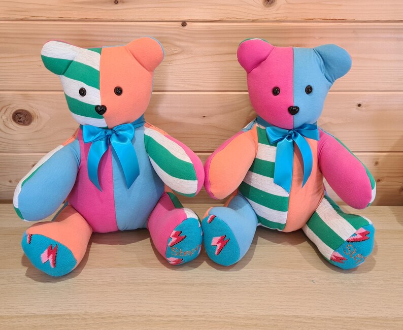 May include: Two colorful handmade teddy bears. One bear is orange, green, blue, and pink with a blue bow. The other bear is pink, blue, orange, and green with a blue bow. Each bear has a lightning bolt design on its foot.
