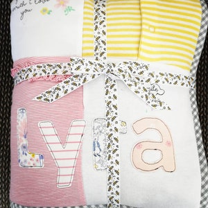 Pode incluir: Um conjunto de presente para bebé embrulhado com uma fita estampada. O conjunto inclui um cobertor branco com o texto "guess how much I love you", um cobertor listrado rosa e branco com o nome "Lyla" e um body listrado amarelo.