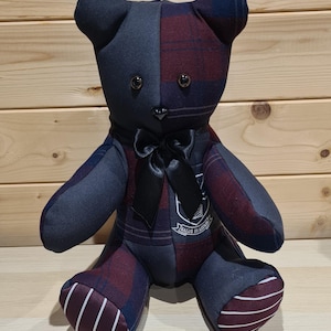 May include: A handmade teddy bear with a patchwork design. The bear is primarily navy blue and burgundy plaid fabric, with a black satin bow. The bear has button eyes and a small emblem on its chest. The bear is sitting on a light wood surface.