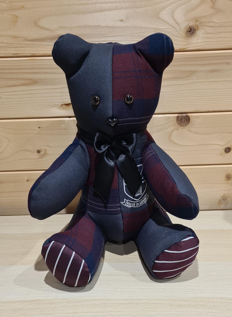 May include: A handmade teddy bear with a patchwork design. The bear is primarily navy blue and burgundy plaid fabric, with a black satin bow. The bear has button eyes and a small emblem on its chest. The bear is sitting on a light wood surface.