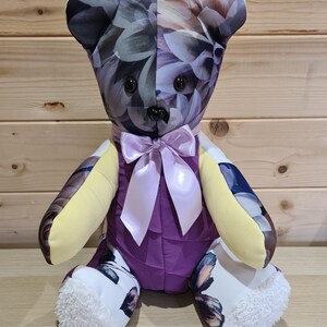 May include: A handmade teddy bear with a floral pattern on the head and arms, and butterfly patterns on the feet. The bear has a purple body, yellow arms, and a light purple bow. The eyes are black.