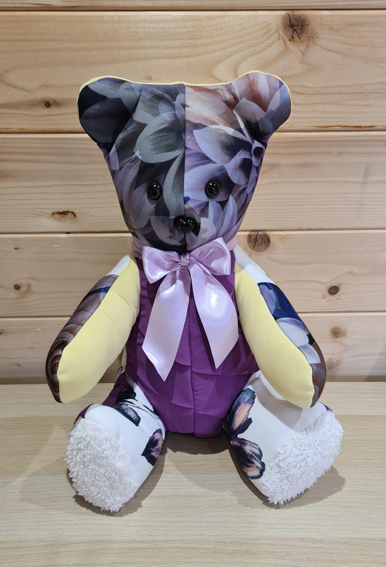 May include: A handmade teddy bear with a floral pattern on the head and arms, and butterfly patterns on the feet. The bear has a purple body, yellow arms, and a light purple bow. The eyes are black.