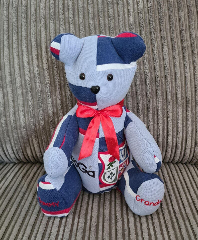 Pu&ograve; includere: Un orsacchiotto a strisce blu e bianche con accenti rossi. L'orso &egrave; fatto di tessuto riutilizzato e ha un fiocco rosso intorno al collo. L'orso ha uno stemma sul petto e le parole "Granda" e "Rugby" sui suoi piedi.