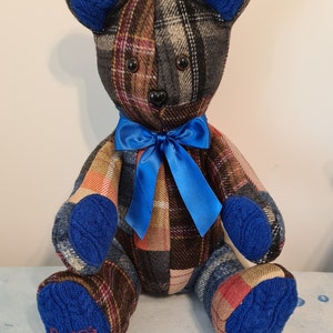 May include: A patchwork plaid teddy bear with blue knitted ears, paws, and a blue satin bow. The bear is sitting on a blue and white patterned surface. The bear's right paw has the name "Pran" embroidered on it.