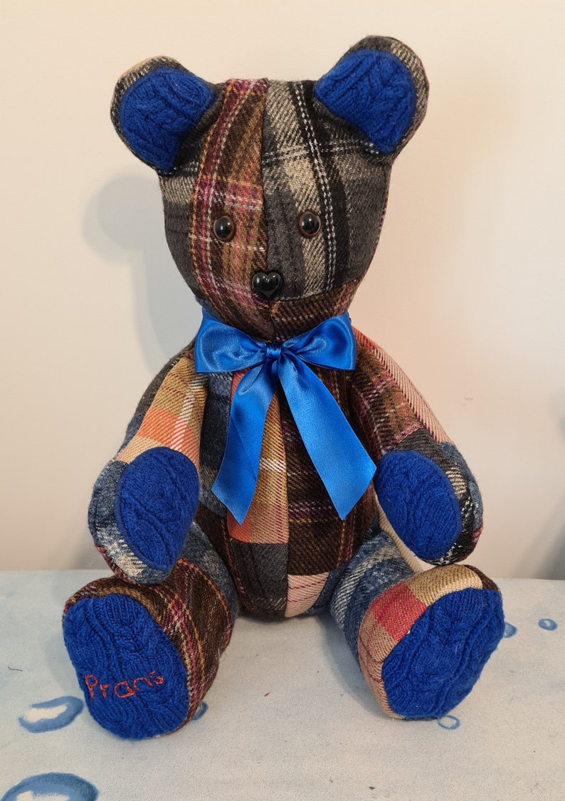 Pu&ograve; includere: Un orsacchiotto di pezza a patchwork a quadri con orecchie, zampe e un fiocco di raso blu a maglia. L'orso &egrave; seduto su una superficie a motivi blu e bianchi. La zampa destra dell'orso ha il nome "Pran" ricamato.