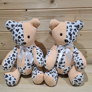 Pu&ograve; includere: Due orsacchiotti di peluche con pelliccia color pesca e cuori neri stampati sul loro tessuto. Gli orsi sono seduti su una superficie di legno.