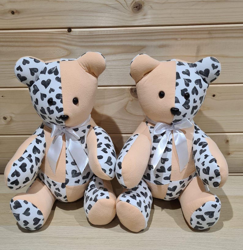 Pu&ograve; includere: Due orsacchiotti di peluche con pelliccia color pesca e cuori neri stampati sul loro tessuto. Gli orsi sono seduti su una superficie di legno.