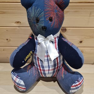May include: A handmade teddy bear with a blue and red firework design on its head. The bear has a white bow tie and a plaid patterned body. The left arm has the text "Lodge's".