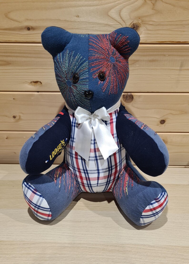 May include: A handmade teddy bear with a blue and red firework design on its head. The bear has a white bow tie and a plaid patterned body. The left arm has the text "Lodge's".