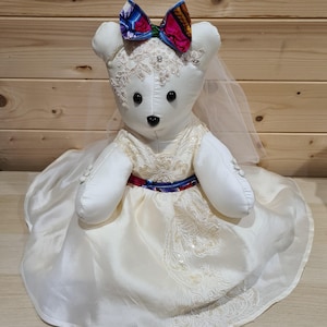 Könnte beinhalten: Ein weißer Teddybär in einem cremefarbenen Hochzeitskleid mit Spitzen- und Pailletten-Details. Der Bär hat eine bunte Schleife, einen Schleier und eine rot-weiß-blaue Schärpe. Der Bär sitzt auf einer hellen Oberfläche.