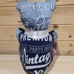 May include: A handmade teddy bear made from repurposed fabric. The bear's head is denim with "PRONE CO 1969" embroidered on it. The body is navy blue with "Vintage" and "Original Parts" printed on it. A white ribbon is tied around the neck.