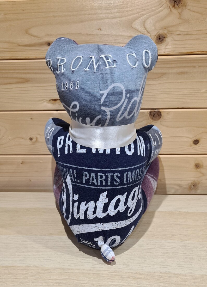 May include: A handmade teddy bear made from repurposed fabric. The bear's head is denim with "PRONE CO 1969" embroidered on it. The body is navy blue with "Vintage" and "Original Parts" printed on it. A white ribbon is tied around the neck.