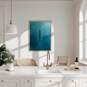 May include: A kitchen scene with a white countertop, sink, and cabinets. A framed blue artwork hangs on the wall. A gold faucet, a bottle, and a plant in a white pot are on the countertop. Two wicker chairs are in the background.