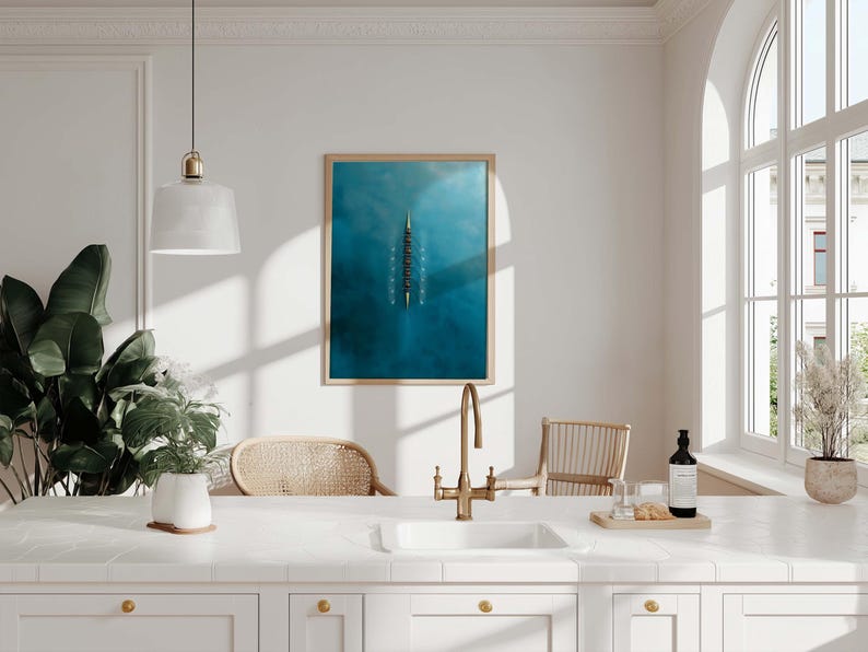 May include: A kitchen scene with a white countertop, sink, and cabinets. A framed blue artwork hangs on the wall. A gold faucet, a bottle, and a plant in a white pot are on the countertop. Two wicker chairs are in the background.