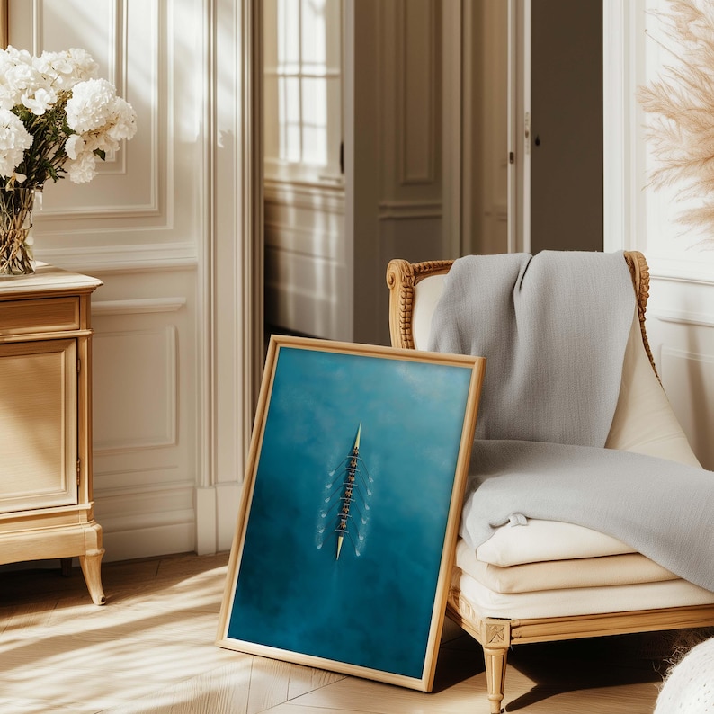 May include: A framed print featuring a top-down view of a rowing team on a teal blue body of water. The wooden frame complements the artwork, which is leaning against a vintage armchair with a gray throw. A vase of white flowers sits on a wooden cabinet.