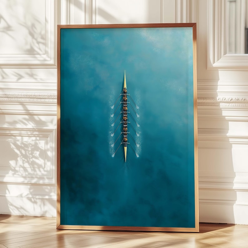 May include: A framed print featuring an aerial view of a rowing team in a yellow boat on turquoise water. The boat is centered, with eight rowers visible. The frame is a light brown color.
