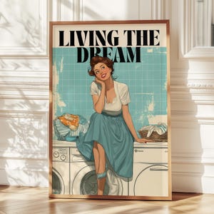 May include: A framed vintage-style print featuring a woman sitting on a washing machine. The print has the text "LIVING THE DREAM" at the top. The woman is wearing a blue skirt and white top, with a basket of laundry nearby. The background is a light blue tile.
