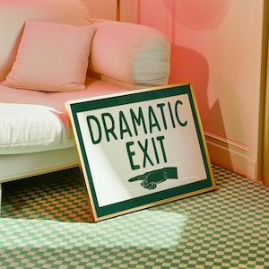 May include: A framed sign with the words "DRAMATIC EXIT" in dark green letters. The sign has a gold frame and a green border. A hand with a pointing finger is also on the sign. The sign is leaning against a white sofa.