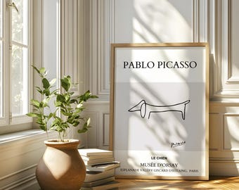 May include: A black and white line drawing of a dog, titled "Le Chien" by Pablo Picasso. The artwork is framed in a light wood frame and displayed on a white wall.