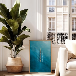 May include: A framed art print featuring a top-down view of a crew team rowing on turquoise water. The artwork is in a wooden frame and is displayed near a window and a large potted plant.