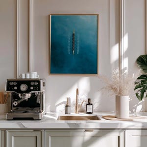 May include: A modern kitchen scene featuring a stainless steel espresso machine on a marble countertop. A framed artwork with a blue and white abstract design hangs above the sink. Decorative elements include a vase with dried flowers and a large potted plant.