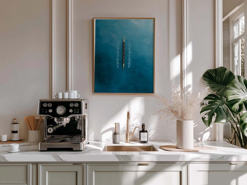 May include: A modern kitchen scene featuring a stainless steel espresso machine on a marble countertop. A framed artwork with a blue and white abstract design hangs above the sink. Decorative elements include a vase with dried flowers and a large potted plant.
